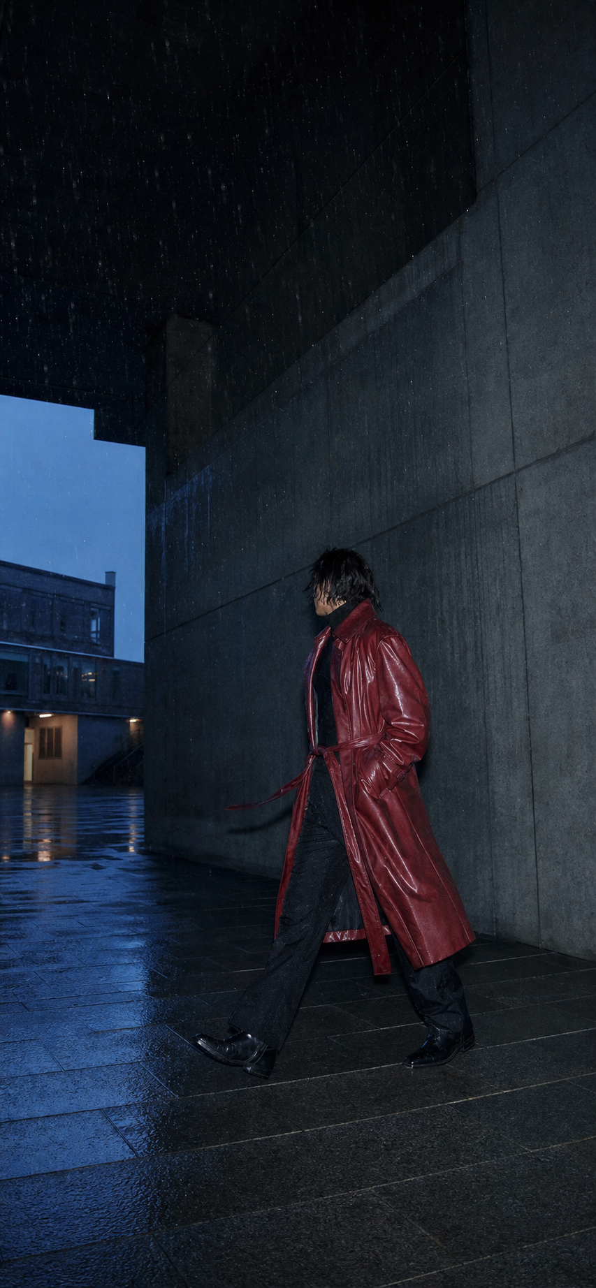 Adult model in a red full-length coat walking through a blue rain-soaked concrete passage